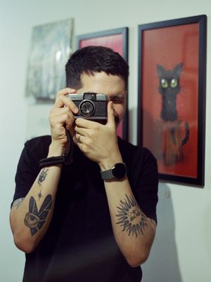 Scanned film photograph of a person (a man with short black hair and a mustache wearing a black t-shirt) looking through the viewfinder of an old camera at the photographer.